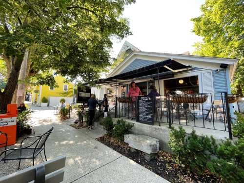 A cozy café with outdoor seating, surrounded by greenery and colorful buildings, on a sunny day.