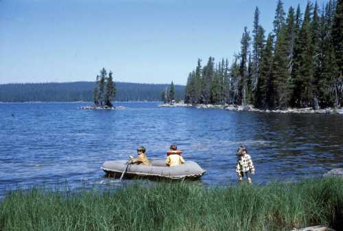 Two children in a small boat on a lake, with a third child walking along the shore, surrounded by trees and mountains.