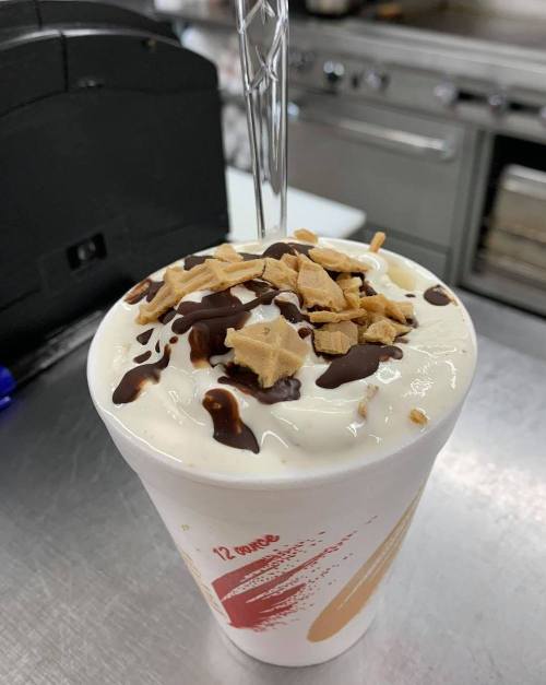 A cup of dessert topped with whipped cream, chocolate drizzle, and crushed cookies, placed on a countertop.