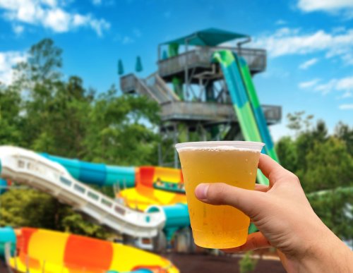 A hand holds a cup of beer in front of colorful water slides and a tall water tower on a sunny day.
