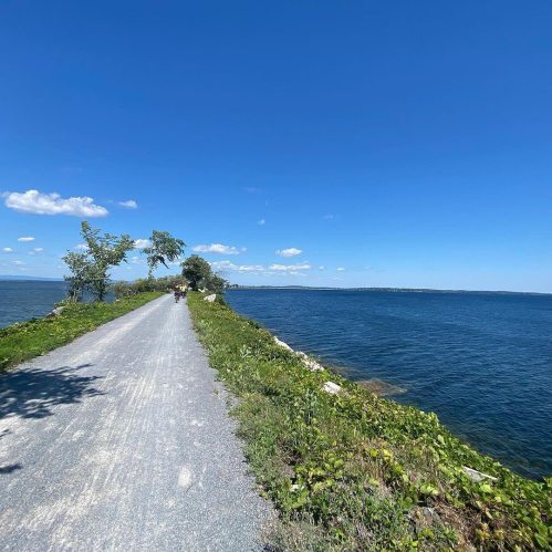 A scenic pathway along a calm blue lake under a clear sky with a few clouds and greenery on the sides.