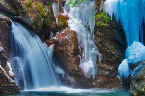 A serene waterfall cascades over rocks, surrounded by icicles and winter foliage, creating a tranquil winter scene.