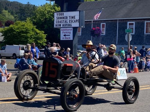 A vintage car in a parade, with two men wearing hats, driving past a crowd and a sign about summer job opportunities.