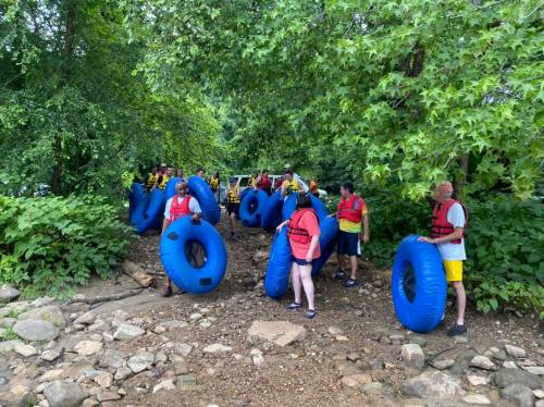 A group of people in life jackets holding blue inner tubes, gathered by a riverbank surrounded by greenery.