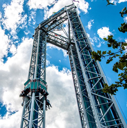 A tall amusement park ride against a blue sky with clouds, featuring riders suspended at the top.