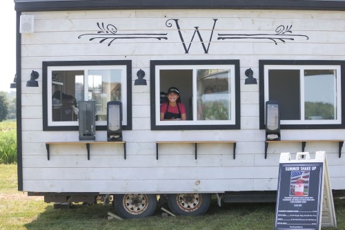A smiling worker stands at a service window of a food truck with a sign promoting summer shakes.