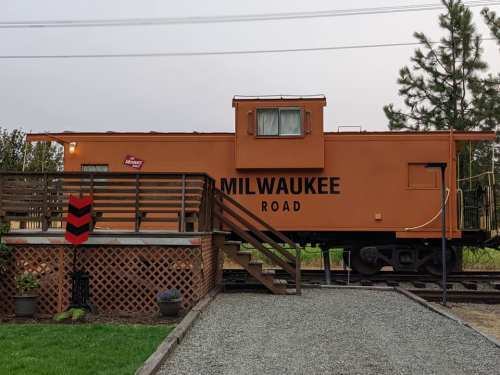 An orange caboose labeled "Milwaukee Road" next to a wooden deck and gravel path, surrounded by trees.