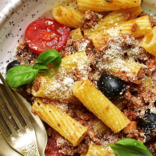 A bowl of rigatoni pasta with ground meat, tomatoes, olives, basil, and grated cheese, served with a fork.