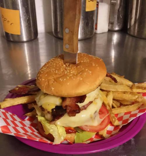A large burger with cheese, bacon, and lettuce, topped with a knife, served with a side of fries on a purple plate.