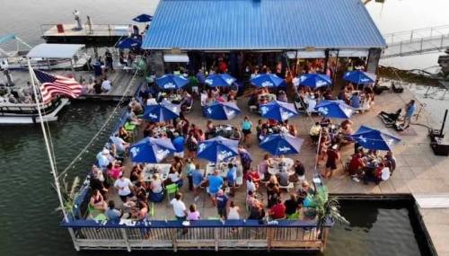 Aerial view of a busy waterfront restaurant with outdoor seating, umbrellas, and a flag, filled with people enjoying their time.