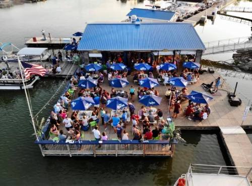 A bustling waterfront restaurant with blue umbrellas, filled with people dining and enjoying the view by the water.