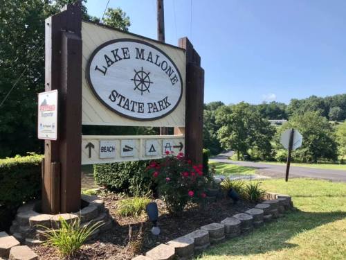 Sign for Lake Malone State Park, featuring directional arrows and surrounded by greenery and flowers.