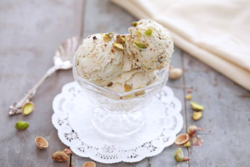 A glass dish holds two scoops of pistachio ice cream, topped with crushed pistachios, on a wooden table.