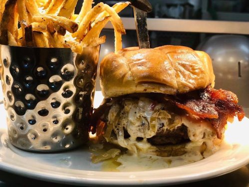 A juicy burger with bacon and melted cheese, served with a side of crispy French fries in a metal cup.