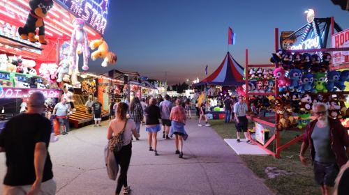 A lively fair scene at dusk, with colorful lights, game booths, and people walking along a bustling pathway.