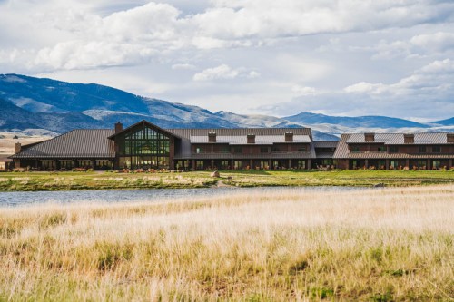 A modern building with large windows sits by a grassy field and a calm lake, surrounded by mountains under a cloudy sky.