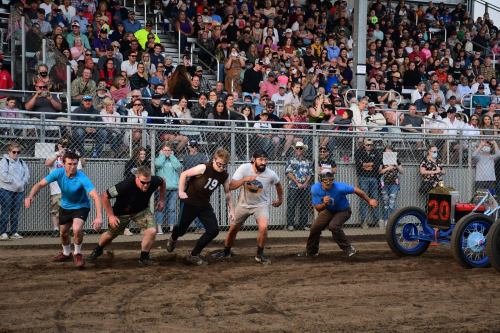 A group of six competitors sprinting in a dirt track race, with a large crowd watching from the stands.