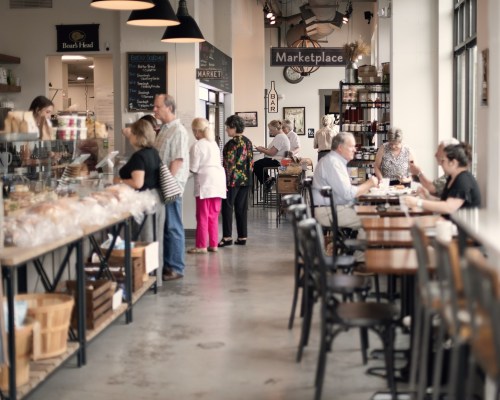 A bustling marketplace with people shopping and dining, featuring a bakery display and various food items.