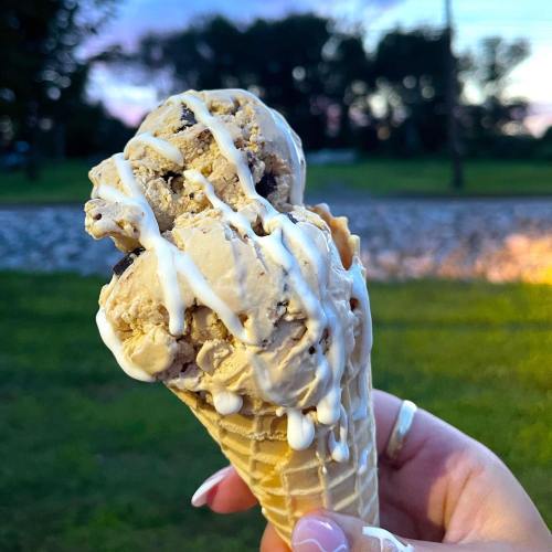 A hand holding a cone with two scoops of ice cream drizzled with white sauce, set against a blurred outdoor background.