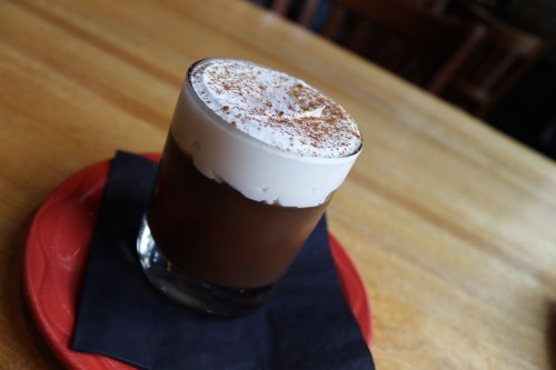 A glass of layered coffee topped with whipped cream and a sprinkle of cocoa, resting on a black napkin on a wooden table.