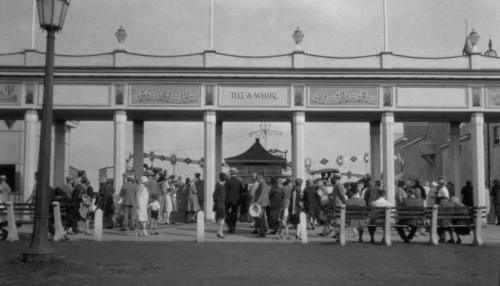 A busy entrance to an amusement park, with crowds of people walking under a decorative archway.