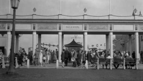 A busy entrance to an amusement park, with crowds of people walking under a decorative archway.