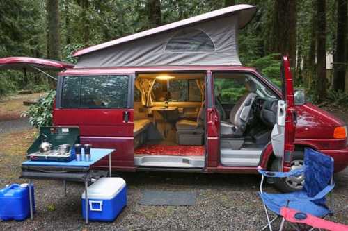 A red camper van parked in a forest, with its side door open, revealing a cozy interior and camping gear outside.