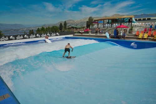 A surfer rides a wave in a man-made surf pool, with spectators and colorful chairs in the background.
