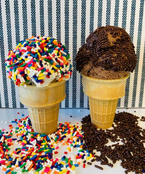 Two ice cream cones: one with colorful sprinkles and the other with chocolate sprinkles, surrounded by more sprinkles.