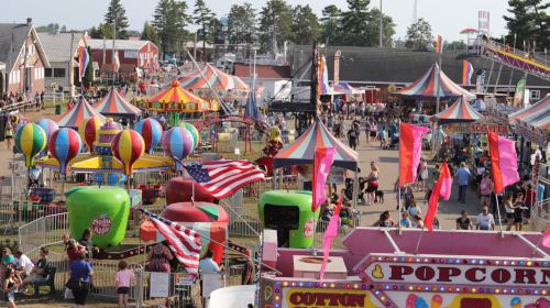 A vibrant fair scene with colorful rides, balloons, and crowds enjoying games and food stalls under a clear sky.