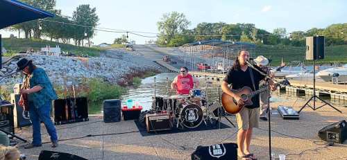 A band performs by the water, with a guitarist, drummer, and bassist near boats and a marina in the background.