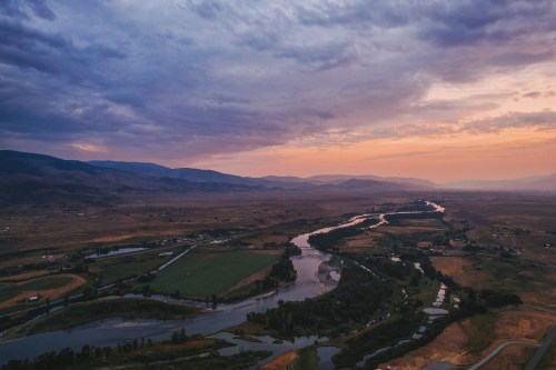 Aerial view of a winding river surrounded by fields and mountains under a colorful sunset sky.