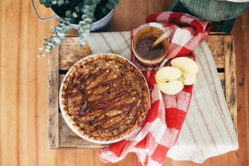 A freshly baked apple pie drizzled with caramel, placed on a checkered cloth with apples and a jar of caramel nearby.