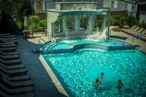 A sunny pool area with a waterfall feature, surrounded by lounge chairs and three people swimming.