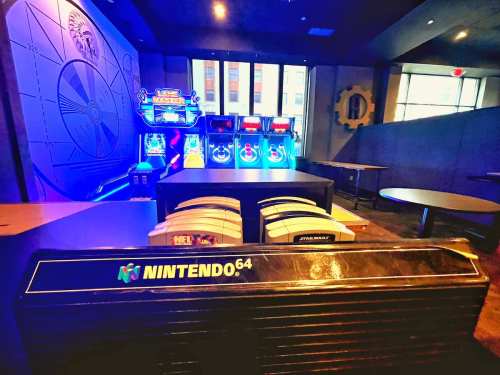 A Nintendo 64 console in the foreground with arcade machines and colorful lights in the background.