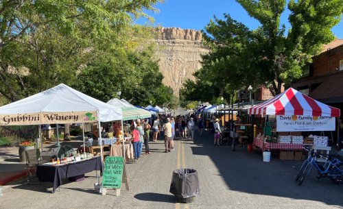 A bustling outdoor market with tents selling goods, set against a backdrop of cliffs and clear blue skies.
