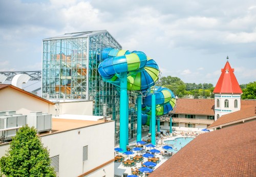 Colorful water slides tower over a pool area, surrounded by lounge chairs and a building with a red roof.