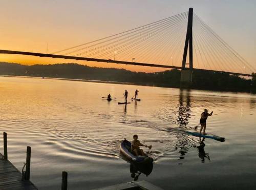 Silhouettes of people paddleboarding and kayaking on a calm river at sunset, with a bridge in the background.