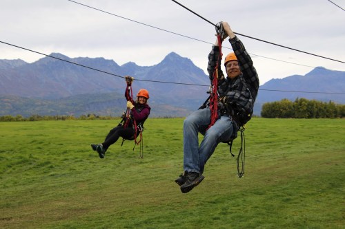 Two people ziplining across a grassy field with mountains in the background, wearing helmets and harnesses.