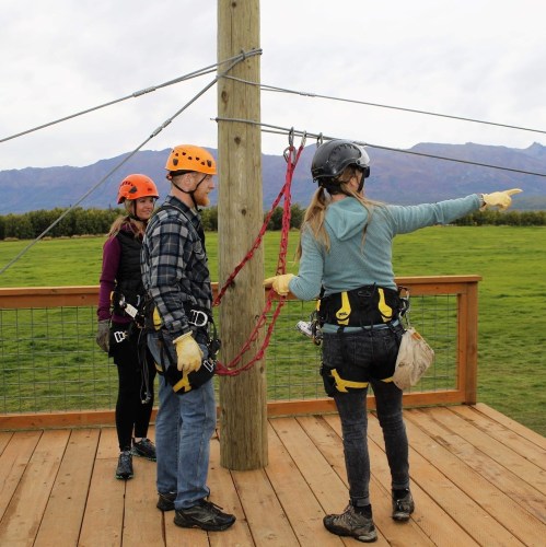 Three people in safety gear stand on a platform, preparing for a zip line activity against a scenic mountain backdrop.