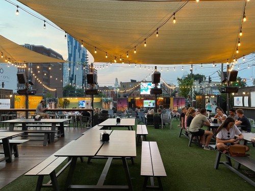 A lively outdoor venue with string lights, tables, and a large screen, filled with people enjoying the atmosphere.