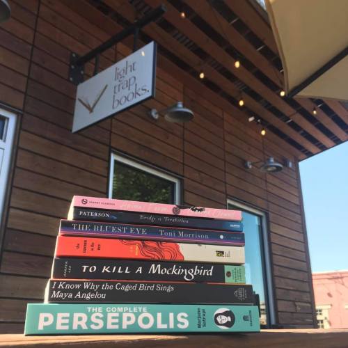 A stack of books in front of a bookstore sign, featuring titles like "To Kill a Mockingbird" and "Persepolis."
