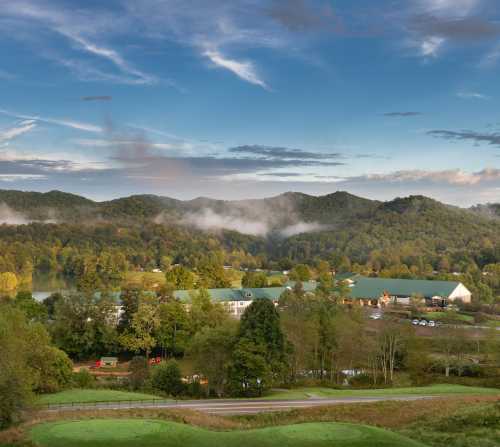 A scenic view of rolling hills and a lake, with mist rising and a building nestled among trees under a blue sky.