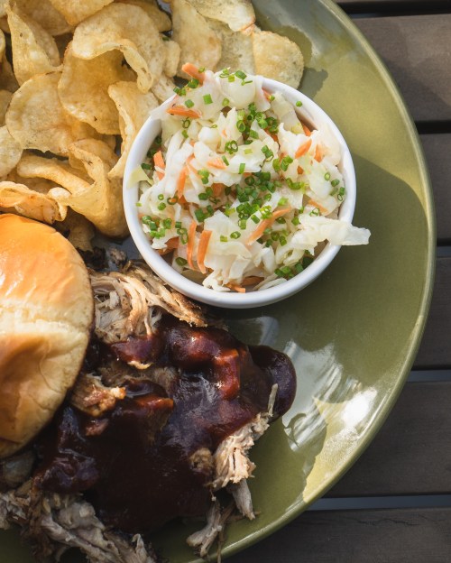 A plate featuring pulled pork with barbecue sauce, a sandwich bun, coleslaw, and potato chips.