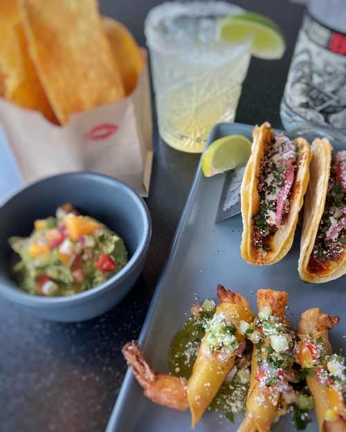 A plate featuring tacos, shrimp, guacamole, tortilla chips, and a margarita with lime.