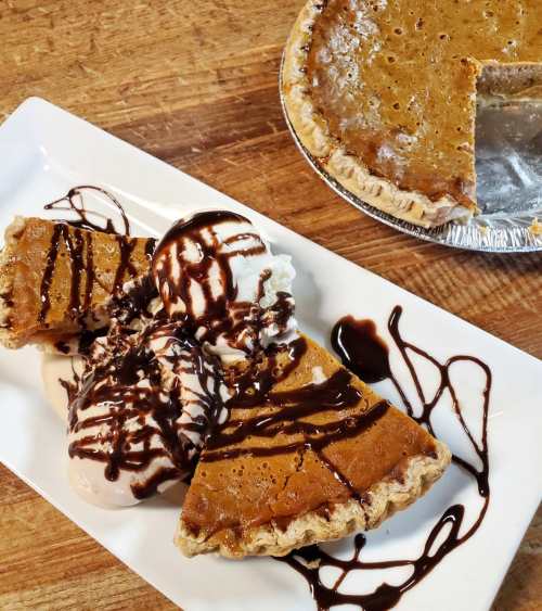 A plate with three slices of pumpkin pie topped with ice cream and chocolate sauce, alongside a pie with a missing slice.