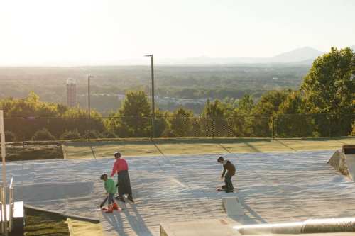 Two children skateboarding on a smooth surface with a scenic view of mountains and a city in the background.