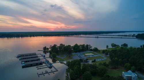 Aerial view of a serene lake at sunset, with docks, trees, and a distant bridge under a colorful sky.