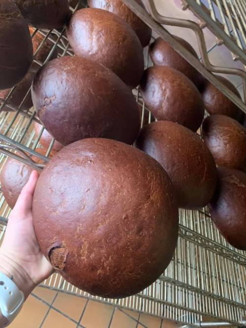 A hand holding a large, round, dark brown bread loaf, with more loaves displayed on a rack in the background.