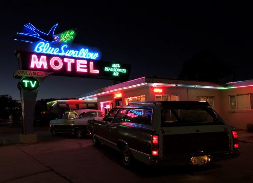 Neon sign of Blue Swallow Motel at night, with classic cars parked outside and colorful lights illuminating the building.